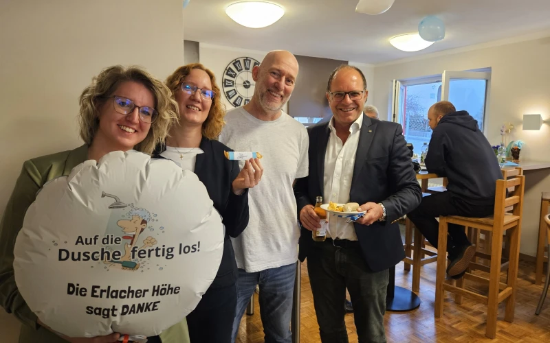 Vier Personen stehen hintereinander in einem Raum, zwei Frauen und zwei M&auml;nner, in lockerer Atmosph&auml;re. Eine Frau h&auml;lt einen runden Lufballon mit der Aufschrift "Auf die Dusche fertig los! Die Erlacher H&ouml;he sagt DANKE". Die Gruppe scheint in einem Bistroraum zu sein, mit Holzm&ouml;beln und Beleuchtung im Hintergrund.