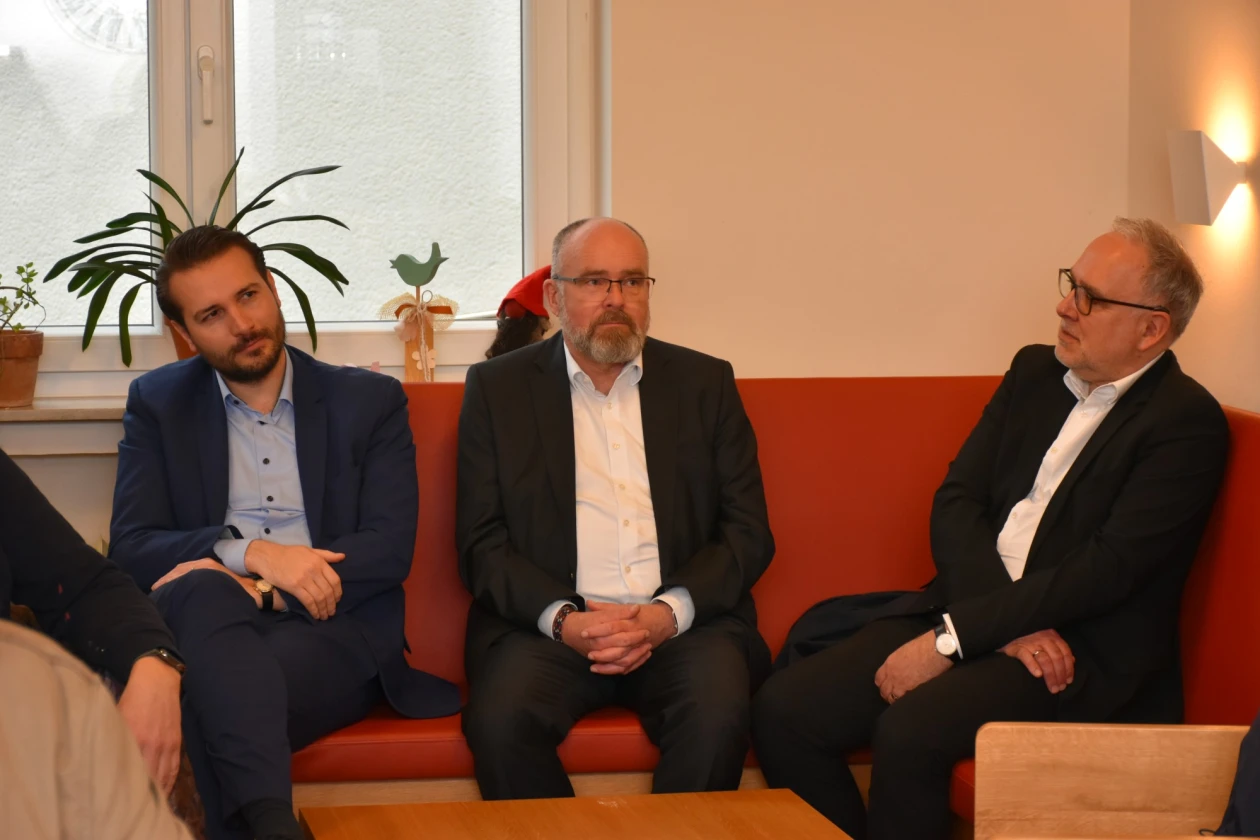 Drei M&auml;nner sitzen auf einem roten Sofa, gekleidet in Business-Anz&uuml;gen. Im Hintergrund stehen Pflanzen an einem Fenster in einem hellen, schlichten Raum. Sie scheinen sich in einem Gespr&auml;ch oder einer Besprechung zu befinden, mit unterschiedlichen K&ouml;rperhaltungen und Blickrichtungen.