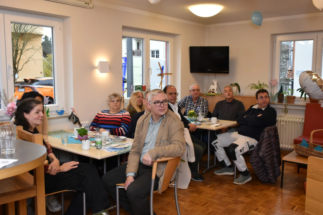 Mehrere Menschen sitzen an Tischen in einem hellen, modernen Raum mit gro&szlig;en Fenstern und einem Fernseher an der Wand. Die Gruppe scheint sich in einer entspannten Umgebung zu treffen, mit verschiedenen Personen in unterschiedlichen Kleidungsst&uuml;cken an eingedeckten Tischen..