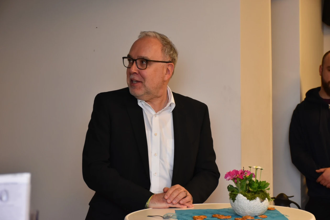 Mann mit Brille und schwarzem Sakko steht an einem Tisch mit Blumen und Serviette, den Blick seitlich gerichtet.