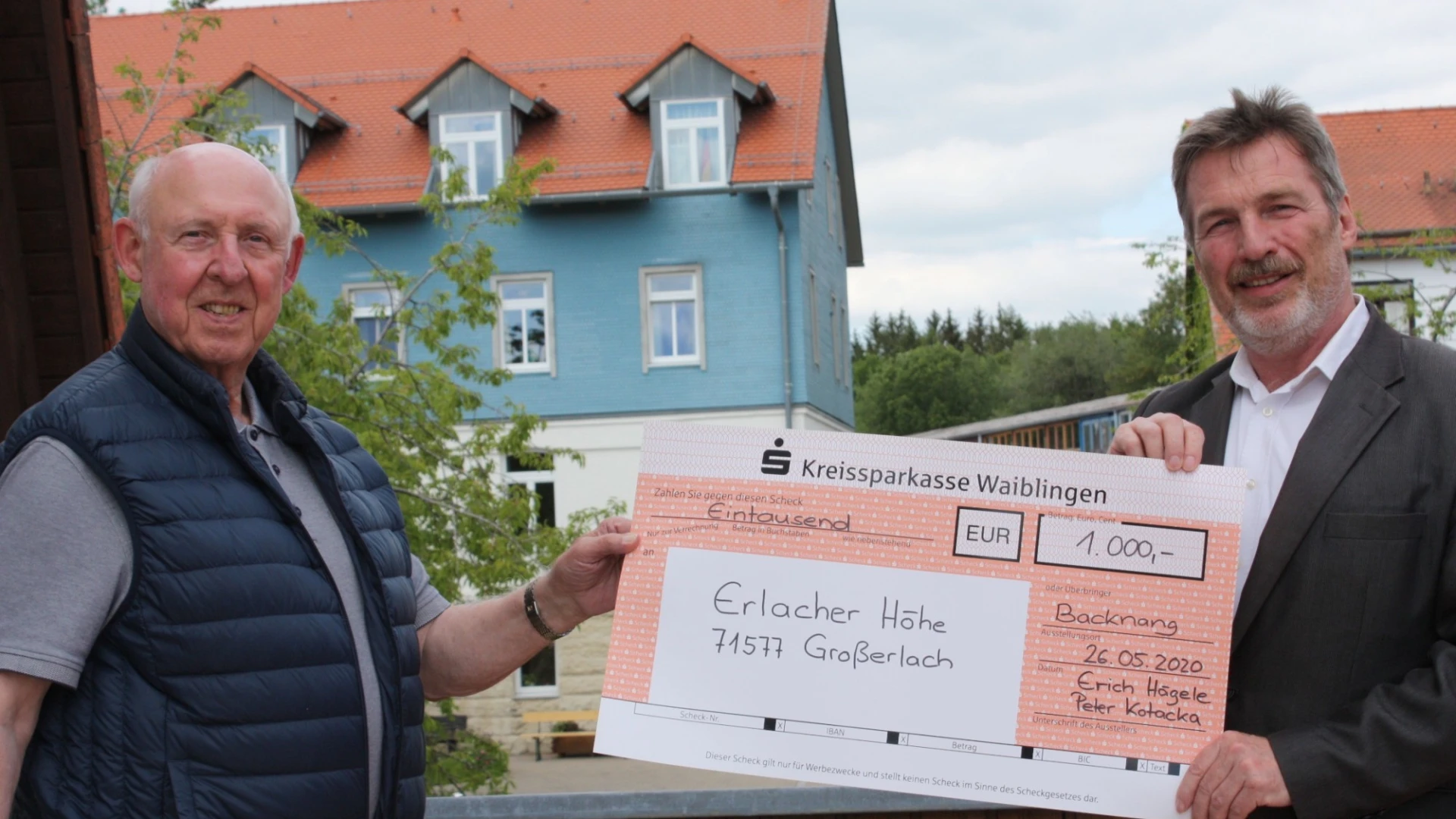 Zwei Personen halten gemeinsam einen Scheck in der Hand. Im Hintergrund sind B&auml;ume und zwei H&auml;user zu sehen.