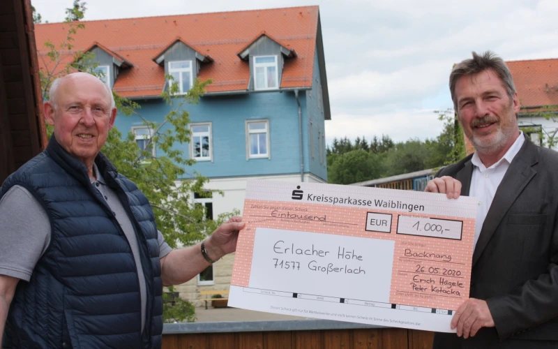 Zwei Personen halten gemeinsam einen Scheck in der Hand. Im Hintergrund sind B&auml;ume und zwei H&auml;user zu sehen.