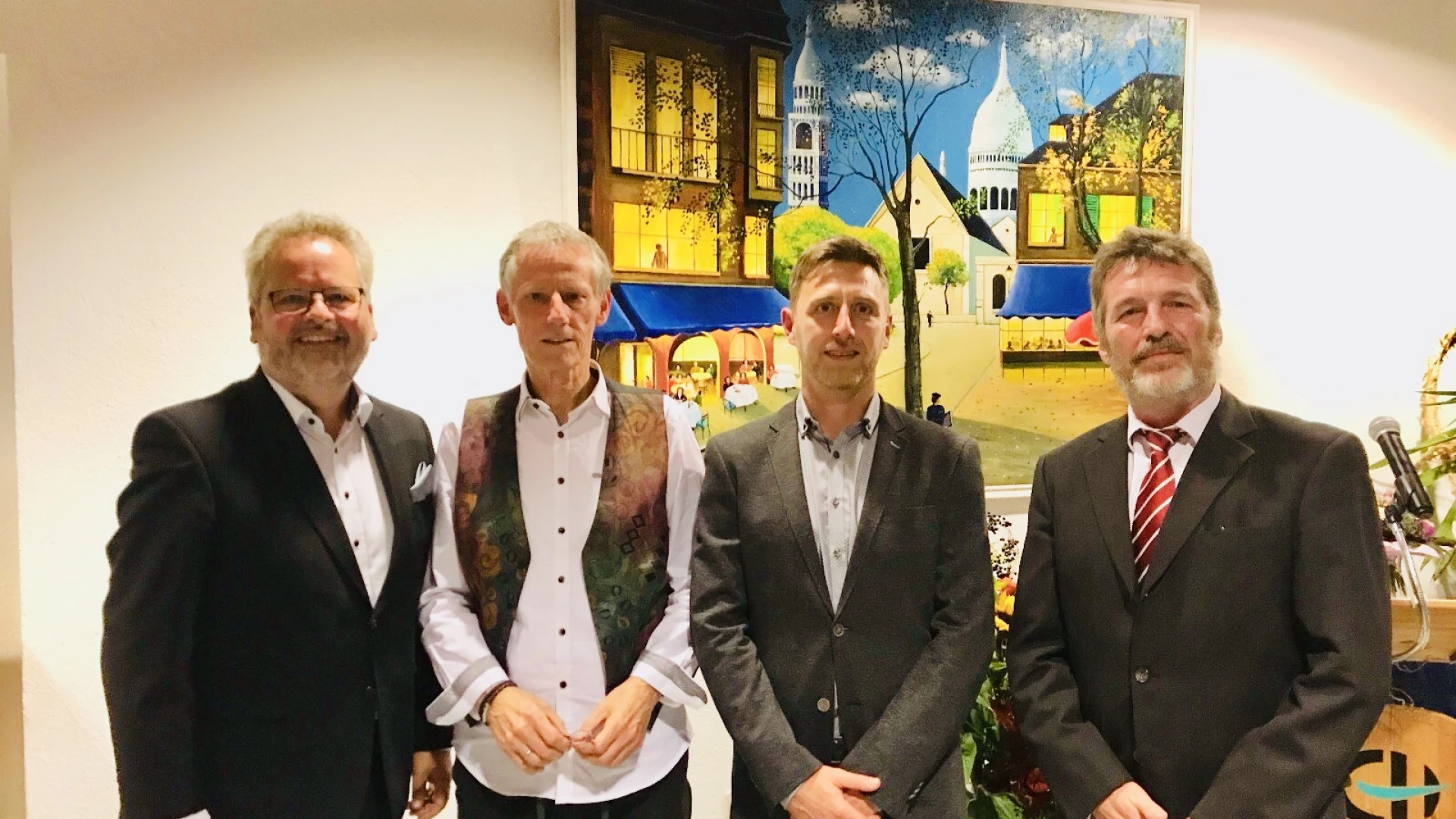 Vier Personen stehen vor einer wei&szlig;en Wand, an der ein buntes Gem&auml;lde h&auml;ngt. Rechts im Hintergrund befindet sich ein Rednerpult mit dem Logo der Erlacher H&ouml;he.