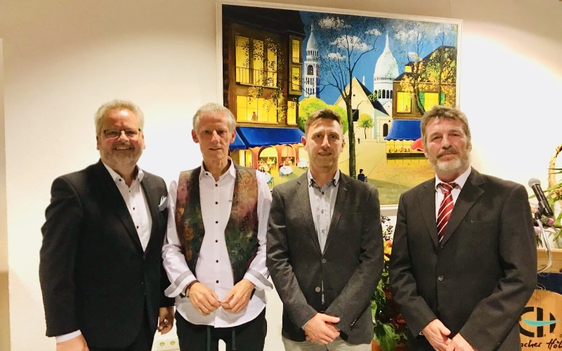 Vier Personen stehen vor einer wei&szlig;en Wand, an der ein buntes Gem&auml;lde h&auml;ngt. Rechts im Hintergrund befindet sich ein Rednerpult mit dem Logo der Erlacher H&ouml;he.