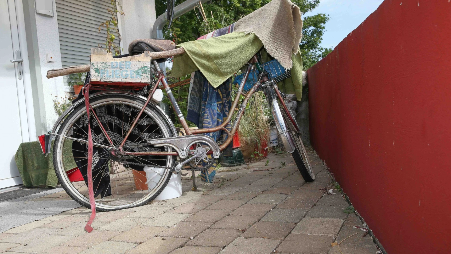 Altes silbernes Fahrrad mit Gep&auml;cktr&auml;ger und plattem Hinterrad, abgestellt auf gepflasterten Boden zwischen einer wei&szlig;en Hauswand und einer roten Wand. Ein Tuch ist &uuml;ber den Fahrradlenker geh&auml;ngt, im Hintergrund sind Pflanzen und eine Rollladenfront zu sehen.