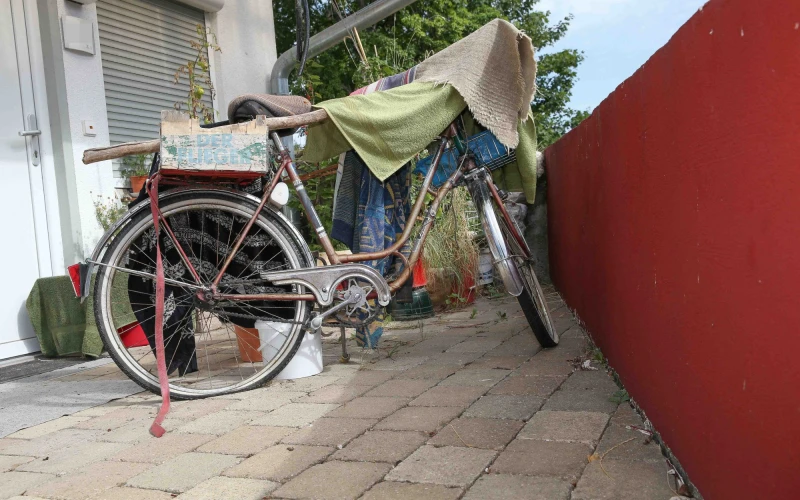 Altes silbernes Fahrrad mit Gep&auml;cktr&auml;ger und plattem Hinterrad, abgestellt auf gepflasterten Boden zwischen einer wei&szlig;en Hauswand und einer roten Wand. Ein Tuch ist &uuml;ber den Fahrradlenker geh&auml;ngt, im Hintergrund sind Pflanzen und eine Rollladenfront zu sehen.