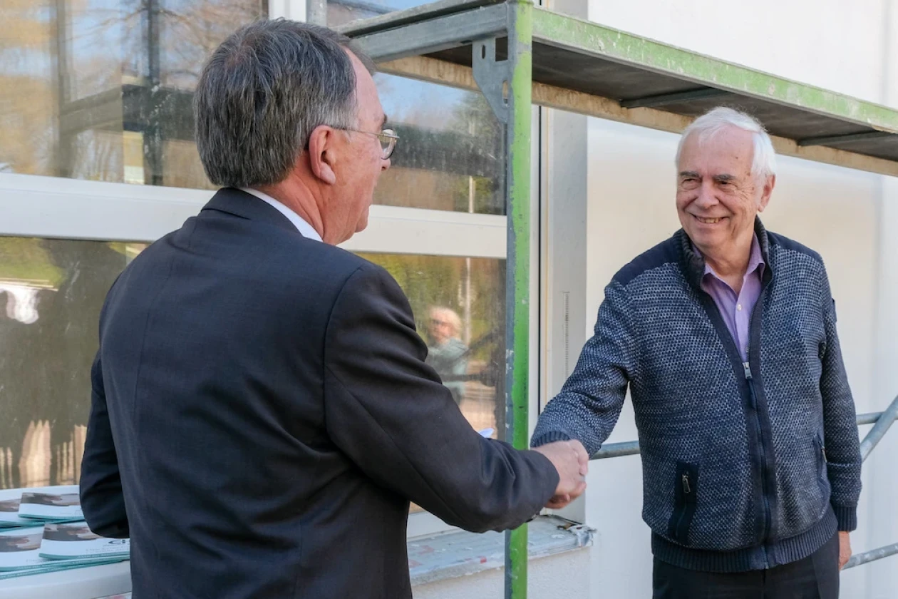 Zwei &auml;ltere M&auml;nner sch&uuml;tteln sich vor einem Geb&auml;ude die Hand, einer tr&auml;gt eine Brille und einen Anzug, der andere einen Pullover und ein Hemd.