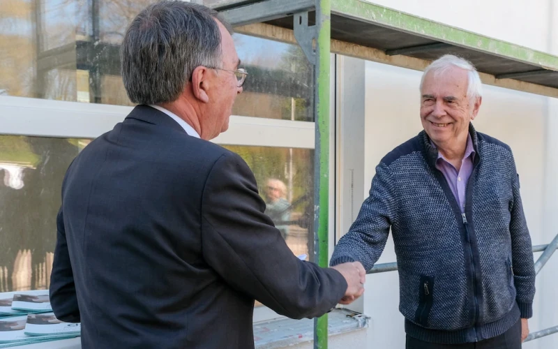 Zwei &auml;ltere M&auml;nner sch&uuml;tteln sich vor einem Geb&auml;ude die Hand, einer tr&auml;gt eine Brille und einen Anzug, der andere einen Pullover und ein Hemd.