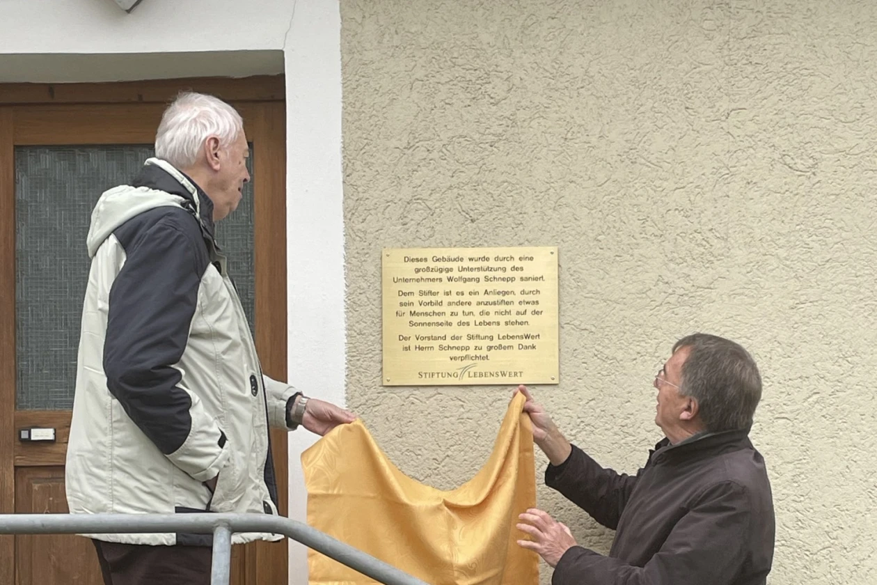 Eine messingfarbene Gedenktafel wird an einem Geb&auml;ude enth&uuml;llt. Zwei M&auml;nner stehen vor dem Geb&auml;ude auf der Eingangstreppe.