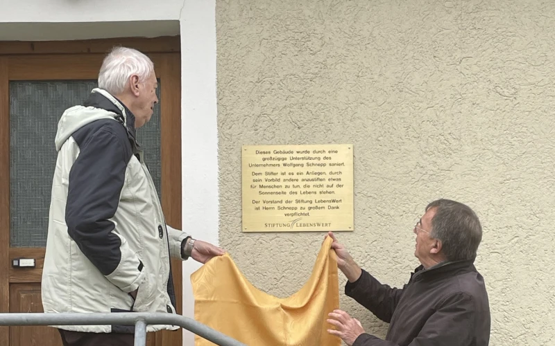 Eine messingfarbene Gedenktafel wird an einem Geb&auml;ude enth&uuml;llt. Zwei M&auml;nner stehen vor dem Geb&auml;ude auf der Eingangstreppe.