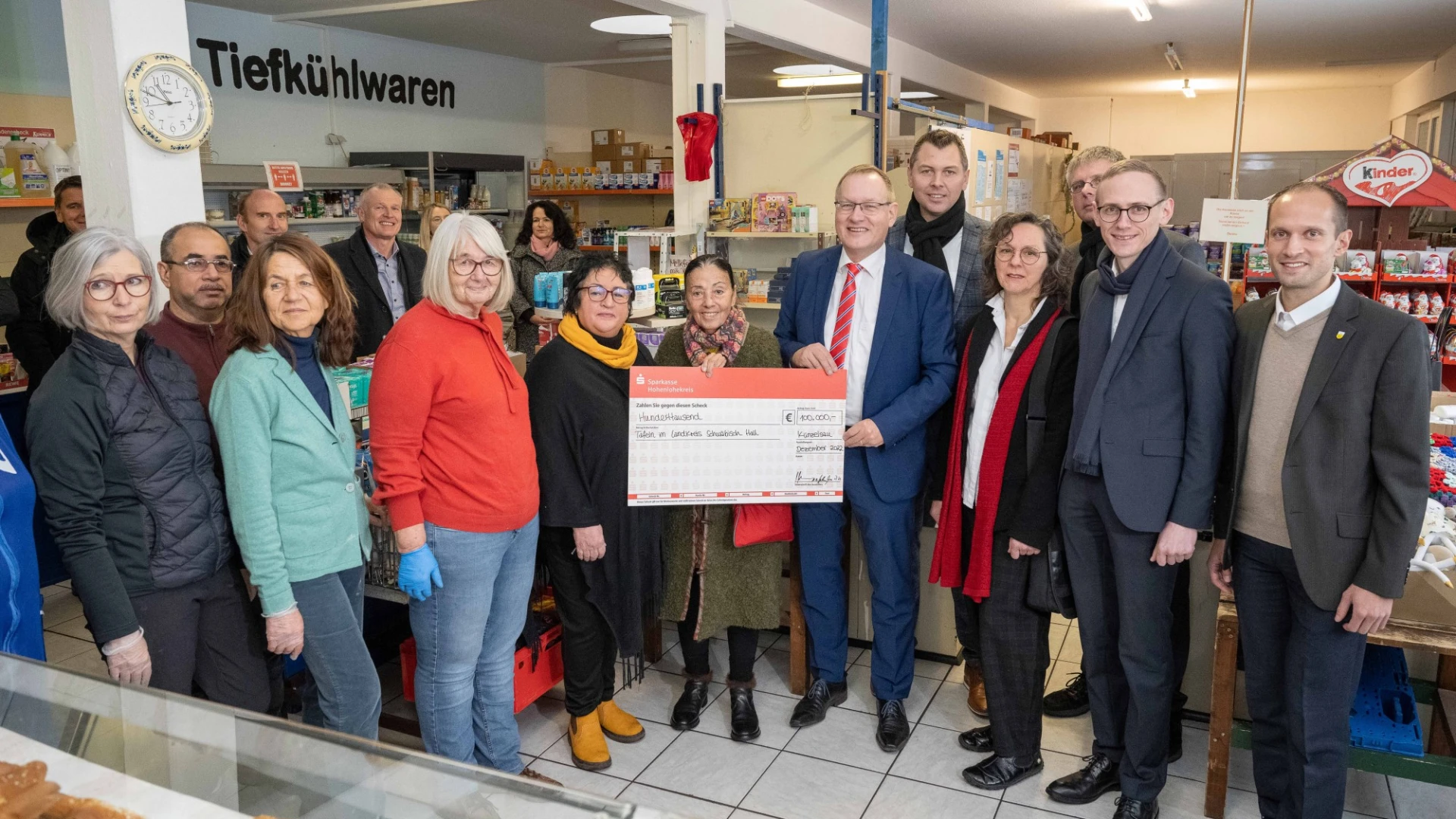 Gruppenfoto in einem Lebensmittelladen mit mehreren Personen, die einen symbolischen Scheck &uuml;bergeben, umgeben von Regalen und Produkten, ein Moment der Zusammenarbeit und Unterst&uuml;tzung.