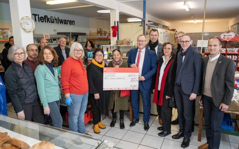 Gruppenfoto in einem Lebensmittelladen mit mehreren Personen, die einen symbolischen Scheck &uuml;bergeben, umgeben von Regalen und Produkten, ein Moment der Zusammenarbeit und Unterst&uuml;tzung.