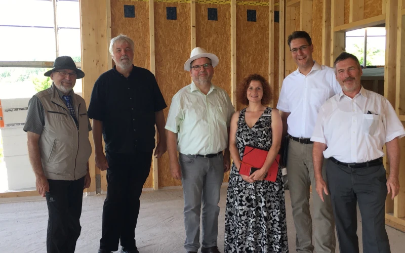 Sechs Menschen in einem Neubau vor Holzst&auml;nderw&auml;nden. Sie blicken in die Kamera und l&auml;cheln.