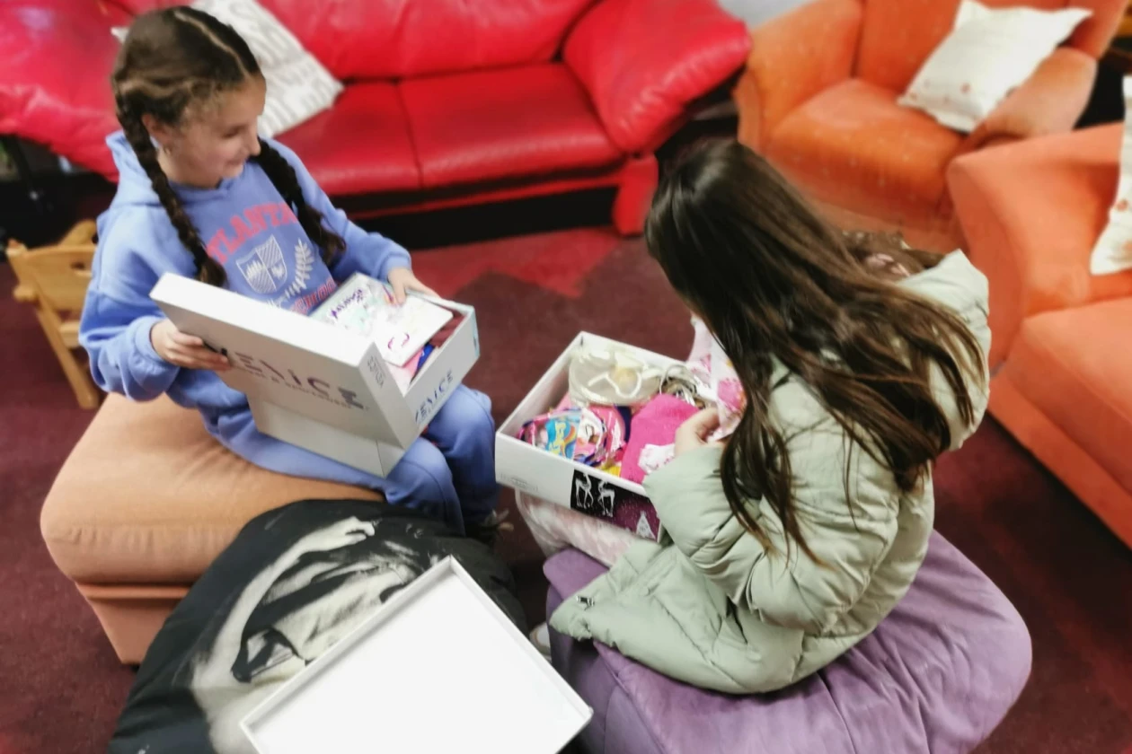 Zwei Kinder sitzen auf einem Sessel und einer Sitzgelegenheit, jeweils mit einer Schachtel in den H&auml;nden. Sie scheinen in einer h&auml;uslichen Umgebung mit roten und orangefarbenen M&ouml;beln konzentriert mit etwas zu besch&auml;ftigt zu sein.