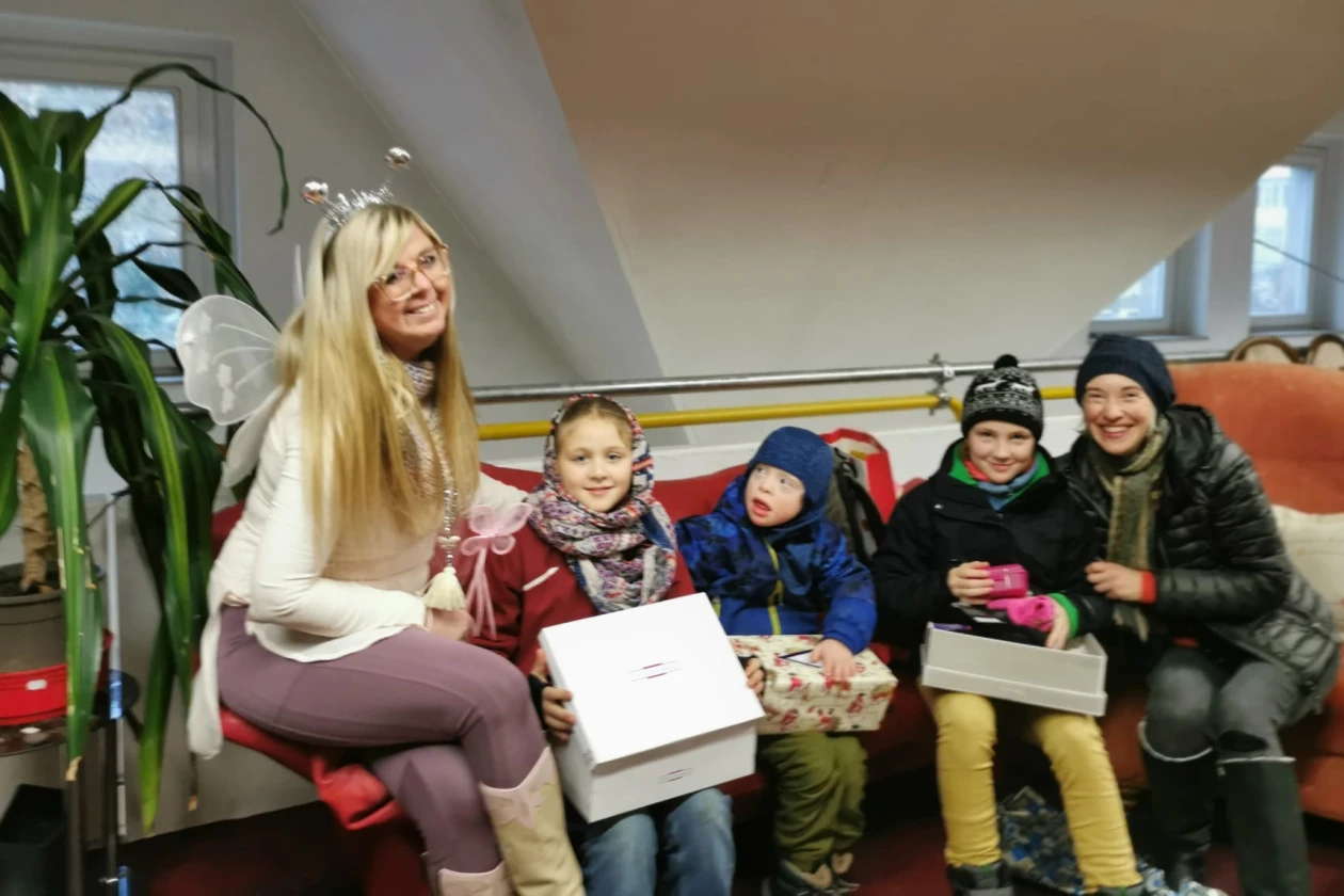 Blonde Frau mit Fl&uuml;geln sitzt mit drei Kindern und einer Frau auf einer Sitzbank, alle tragen Winterkleidung und halten Geschenkboxen, eine gem&uuml;tliche Indoor-Szenerie mit Zimmerpflanze im Hintergrund.