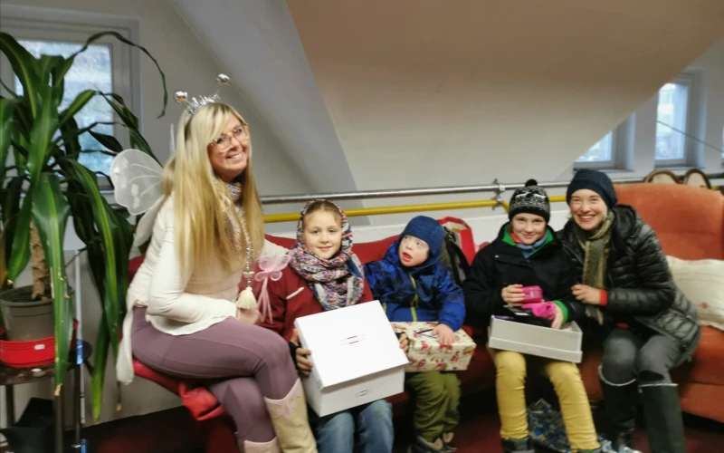 Blonde Frau mit Fl&uuml;geln sitzt mit drei Kindern und einer Frau auf einer Sitzbank, alle tragen Winterkleidung und halten Geschenkboxen, eine gem&uuml;tliche Indoor-Szenerie mit Zimmerpflanze im Hintergrund.