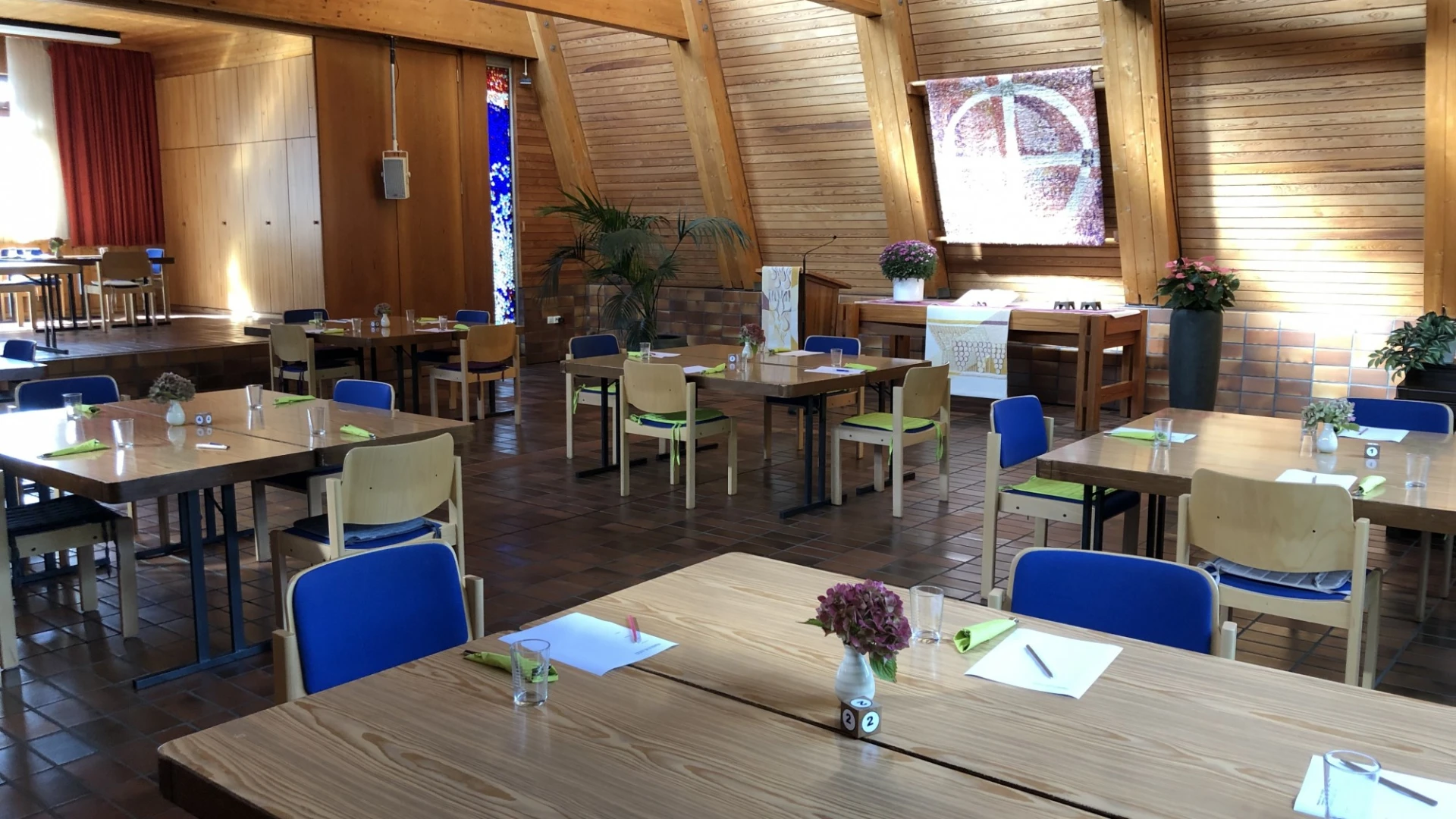 Ein Speisesaal mit mehreren eingedeckten Holztischen und St&uuml;hlen. Auf den Tischen stehen kleine Blumenstr&auml;u&szlig;e, Gl&auml;ser und Besteck mit Servietten.