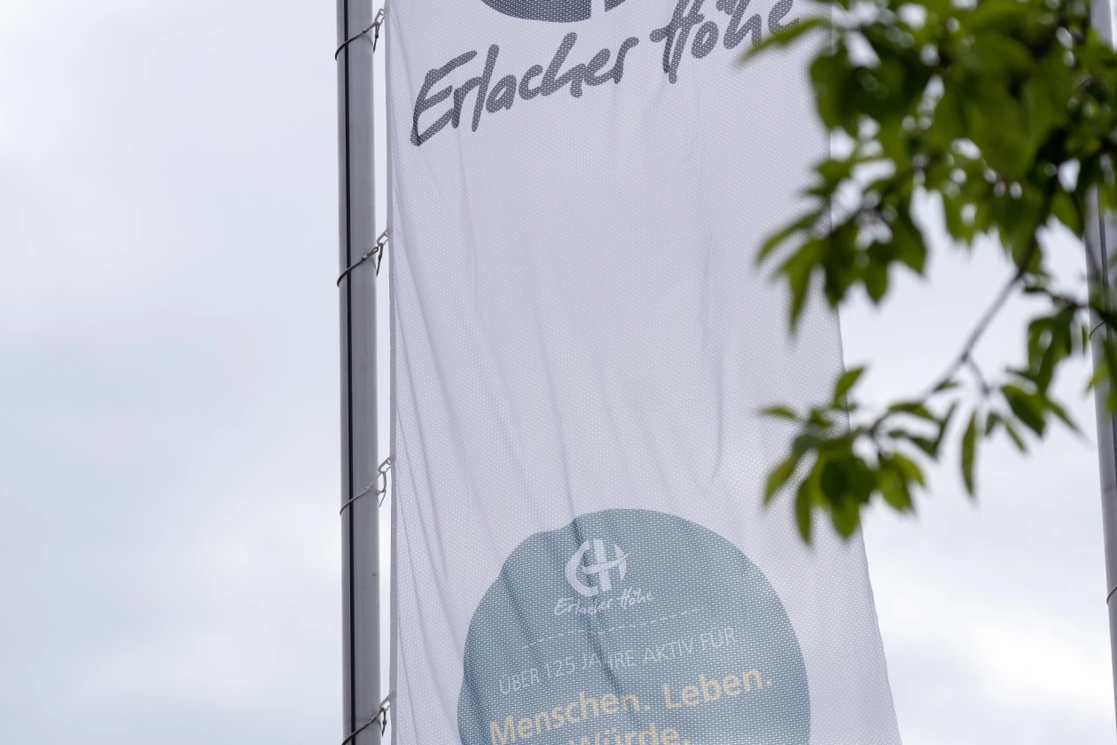 Wei&szlig;e Fahne mit dem Logo und Schriftzug der Erlacher H&ouml;he, vor gr&uuml;nen Bl&auml;ttern und einem bew&ouml;lkten Himmel. Die Flagge zeigt die Webadresse www.erlacher-hoehe.de und einen Kreis mit dem Slogan "Menschen. Leben. W&uuml;rde".