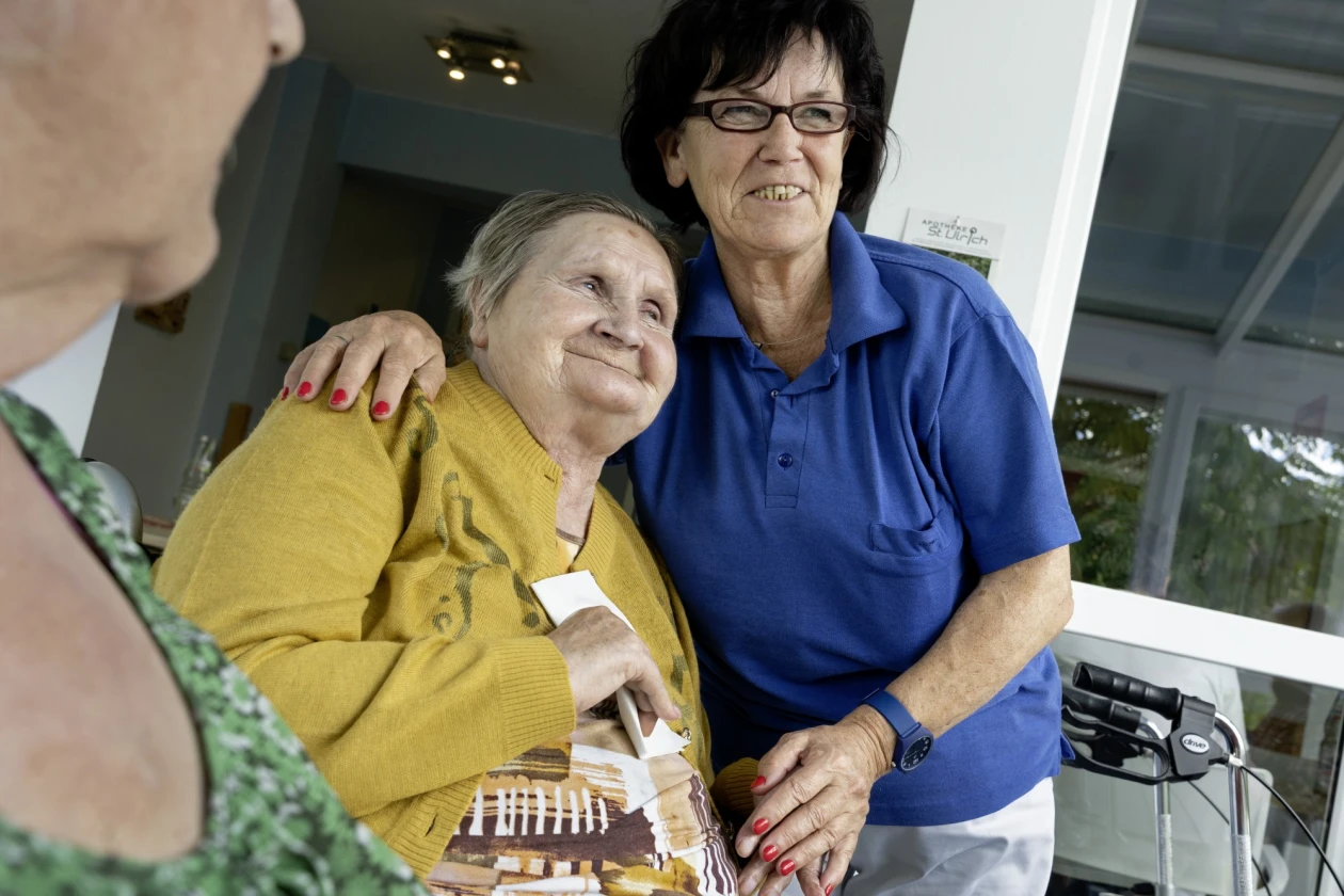 Zwei Frauen l&auml;cheln gemeinsam in die Kamera. Eine tr&auml;gt ein gelbes Oberteil und h&auml;lt ein Taschentuch in der Hand, die andere tr&auml;gt ein blaues T-Shirt und eine Brille. Die Frau im blauen T-Shirt hat ihren Arm um die Schulter der Frau gelegt. Im Vordergrund ist eine weitere Person zu sehen und im Hintergrund steht ein Rollator.