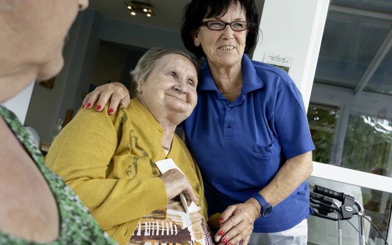 Zwei Frauen l&auml;cheln gemeinsam in die Kamera. Eine tr&auml;gt ein gelbes Oberteil und h&auml;lt ein Taschentuch in der Hand, die andere tr&auml;gt ein blaues T-Shirt und eine Brille. Die Frau im blauen T-Shirt hat ihren Arm um die Schulter der Frau gelegt. Im Vordergrund ist eine weitere Person zu sehen und im Hintergrund steht ein Rollator.