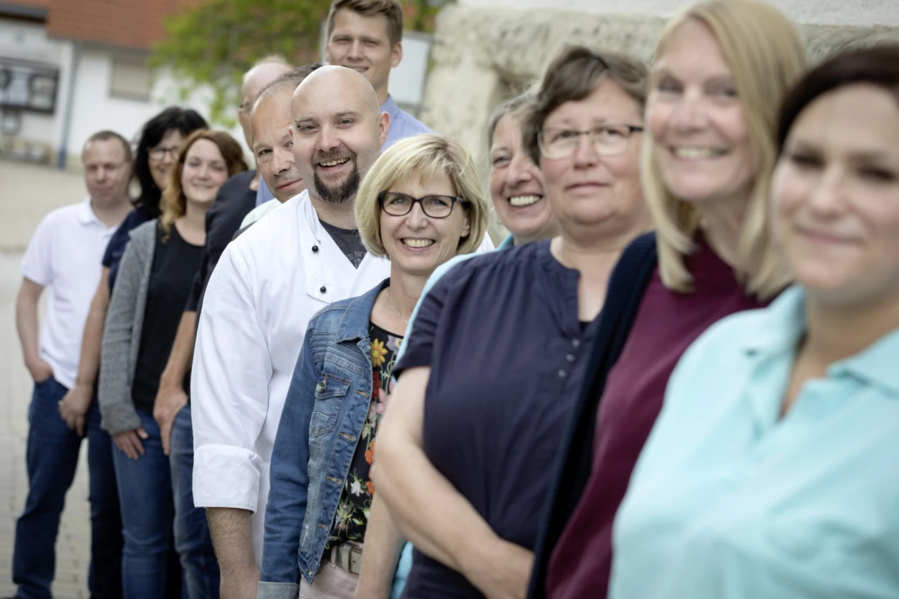 Eine Gruppe von l&auml;chelnden Personen in verschiedenen Kleidungsst&uuml;cken steht hintereinander an einer Wand. Im Hintergrund ist ein Geb&auml;ude und ein Baum zu sehen.