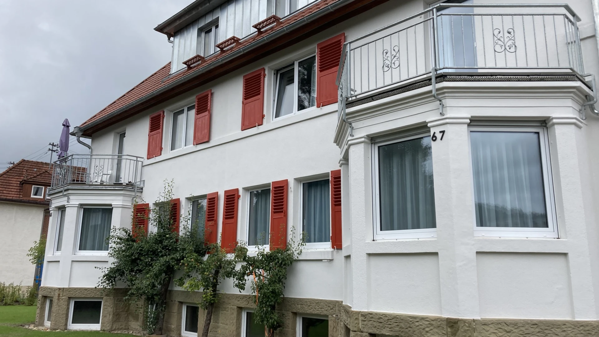 Ein mehrst&ouml;ckiges Geb&auml;ude mit wei&szlig;er Fassade und roten Fensterl&auml;den, sowie zwei Balkonen ist zu sehen.