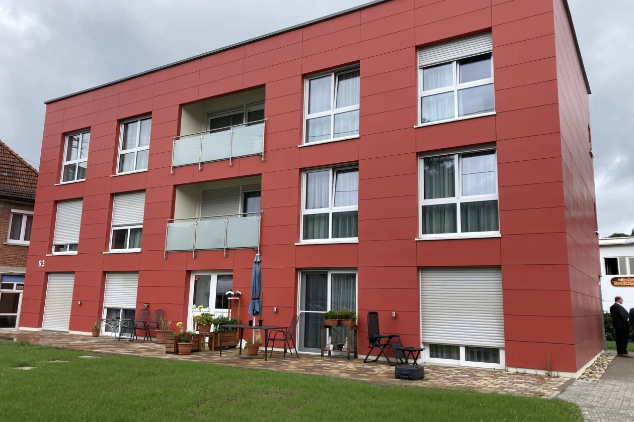 Ein gro&szlig;es rotes Geb&auml;ude mit zwei Balkonen und mehreren Fenstern ist zu sehen. Auf der Terrasse stehen St&uuml;hle, ein Tisch und mehrere Blument&ouml;pfe.