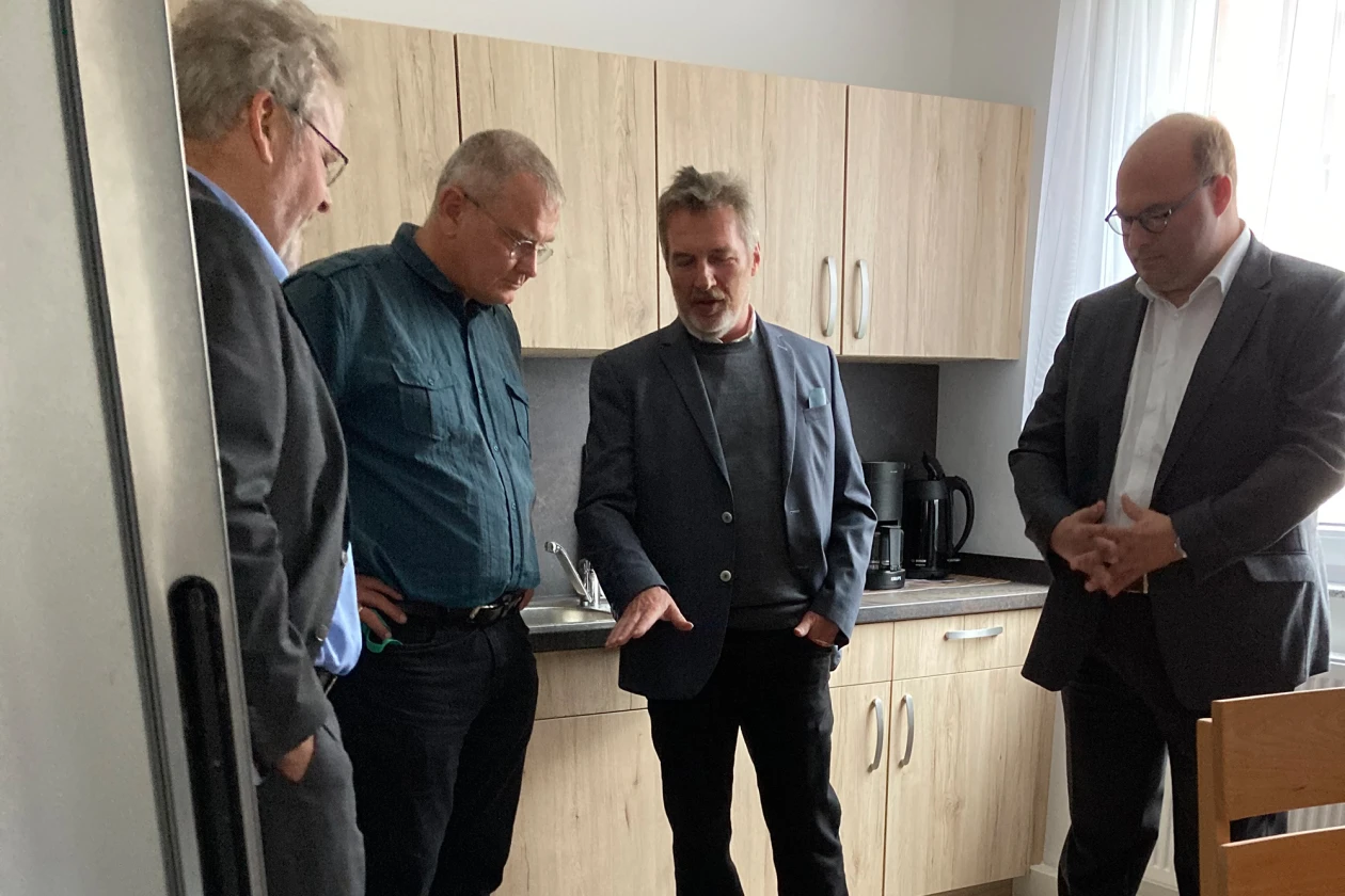 Vier Personen stehen in einer K&uuml;che und blicken auf den Boden, w&auml;hrend ein Mann erz&auml;hlt. Im Hintergrund ist ein Fenster mit wei&szlig;em Vorhang zu sehen.