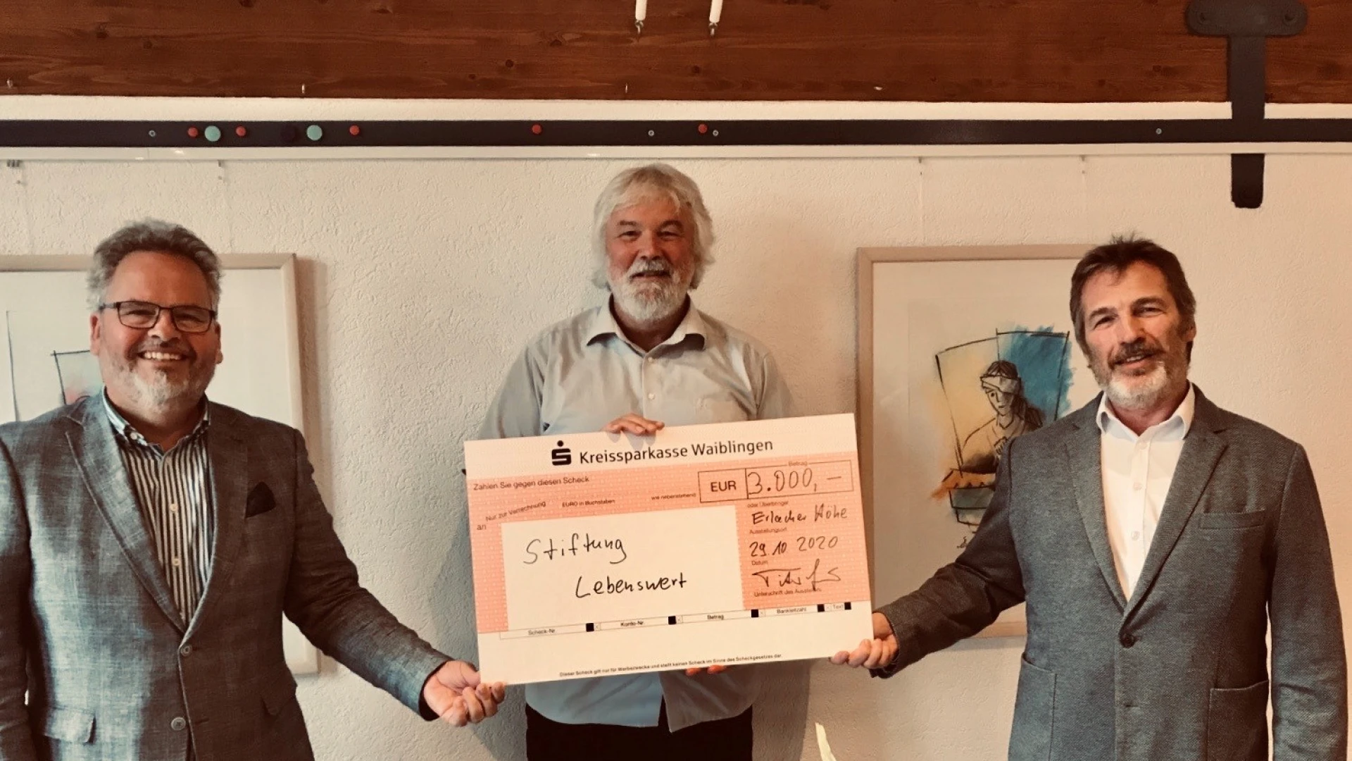 Drei M&auml;nner halten gemeinsam einen Scheck &uuml;ber 3.000 &euro; an die Stiftung Lebenswert in der Hand. An der wei&szlig;en Wand im Hintergrund sind Bilderrahmen zu sehen und im Vordergrund ein Blumenstrau&szlig;.