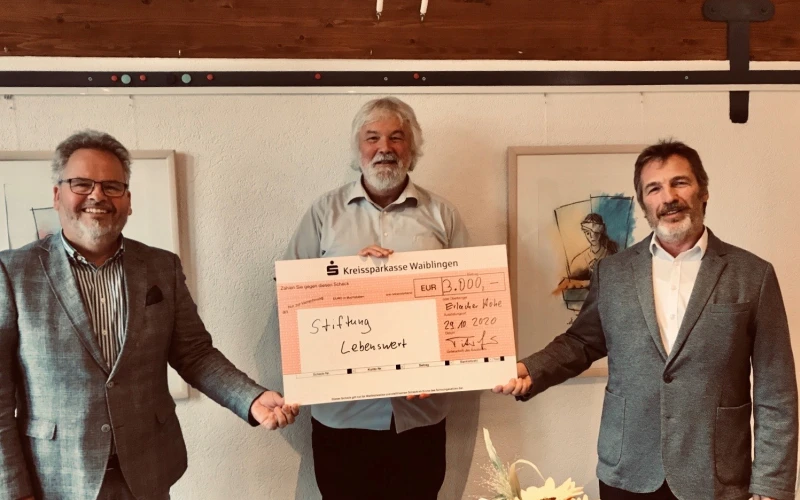 Drei M&auml;nner halten gemeinsam einen Scheck &uuml;ber 3.000 &euro; an die Stiftung Lebenswert in der Hand. An der wei&szlig;en Wand im Hintergrund sind Bilderrahmen zu sehen und im Vordergrund ein Blumenstrau&szlig;.