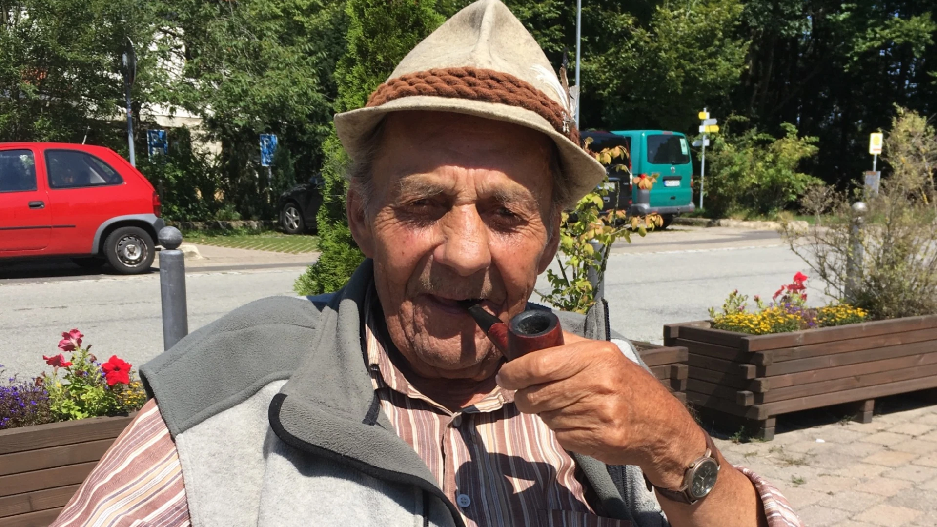 Ein &auml;lterer Mann mit Hut und Pfeife sitzt auf einem Holzstuhl im Freien. Er tr&auml;gt ein gestreiftes Hemd, eine graue Weste und gr&uuml;ne Cordhose. Im Hintergrund sind Blumen, B&auml;ume und ein rotes Auto zu sehen.