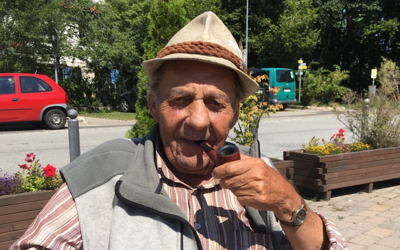 Ein &auml;lterer Mann mit Hut und Pfeife sitzt auf einem Holzstuhl im Freien. Er tr&auml;gt ein gestreiftes Hemd, eine graue Weste und gr&uuml;ne Cordhose. Im Hintergrund sind Blumen, B&auml;ume und ein rotes Auto zu sehen.