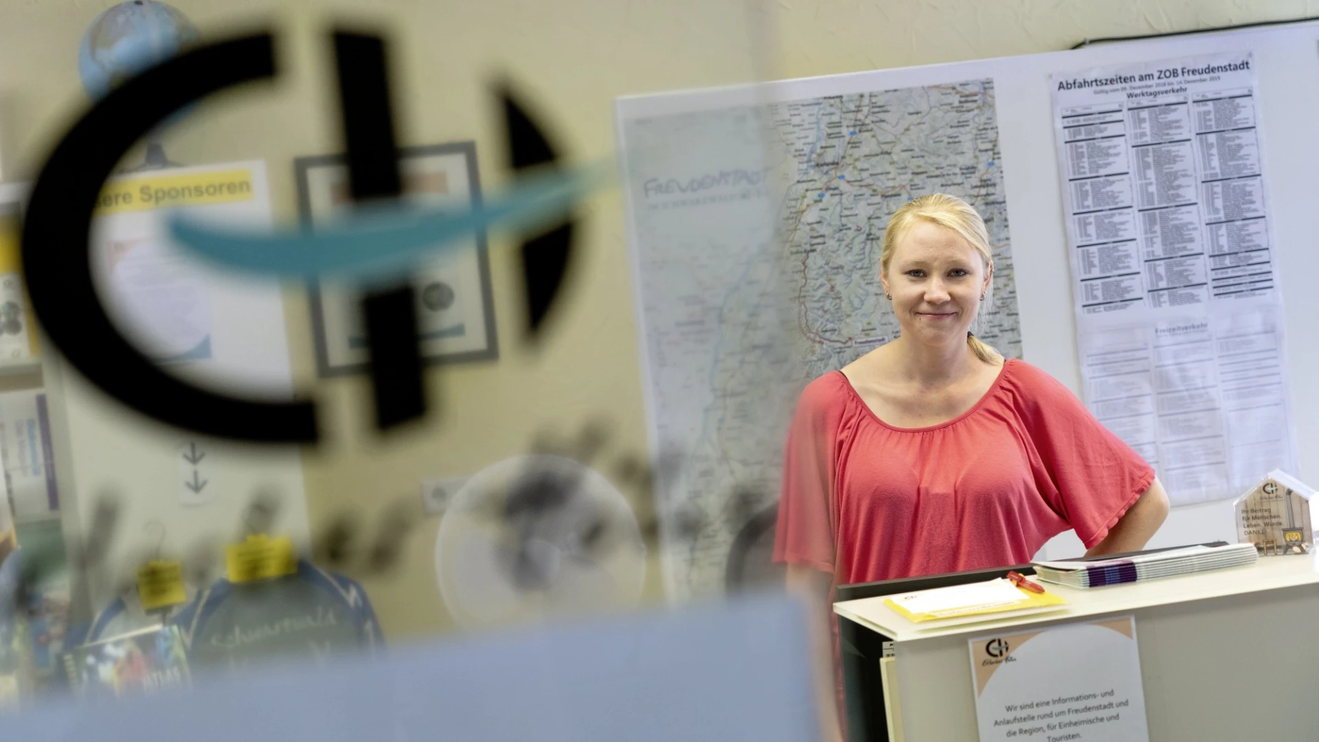 Eine junge, blondhaarige Frau in hellrotem Oberteil steht an einer wei&szlig;en Verkaufstheke vor einer Landkarte und einem Fahrplan. Sie l&auml;chelt freundlich in die Kamera. Im Vordergrund ist unscharf eine Glast&uuml;re mit einem Logo der Erlacher H&ouml;he zu sehen.