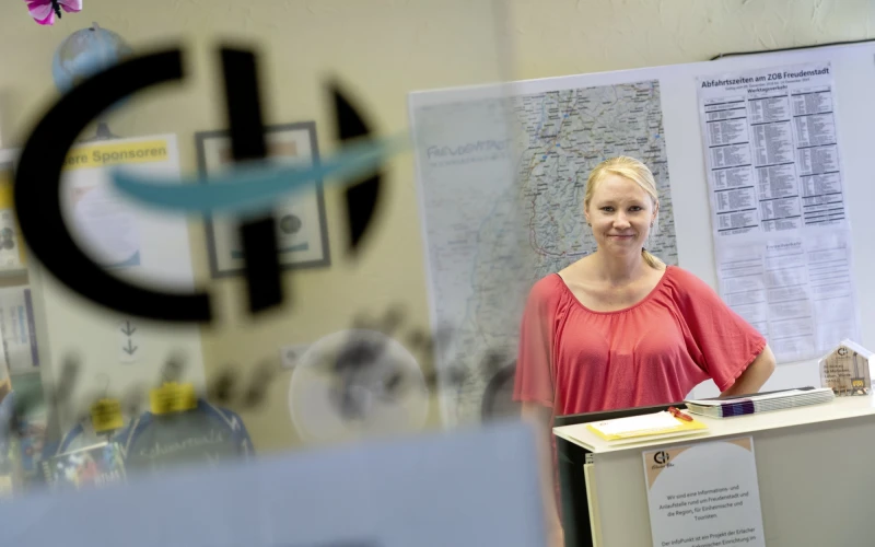 Eine junge, blondhaarige Frau in hellrotem Oberteil steht an einer wei&szlig;en Verkaufstheke vor einer Landkarte und einem Fahrplan. Sie l&auml;chelt freundlich in die Kamera. Im Vordergrund ist unscharf eine Glast&uuml;re mit einem Logo der Erlacher H&ouml;he zu sehen.