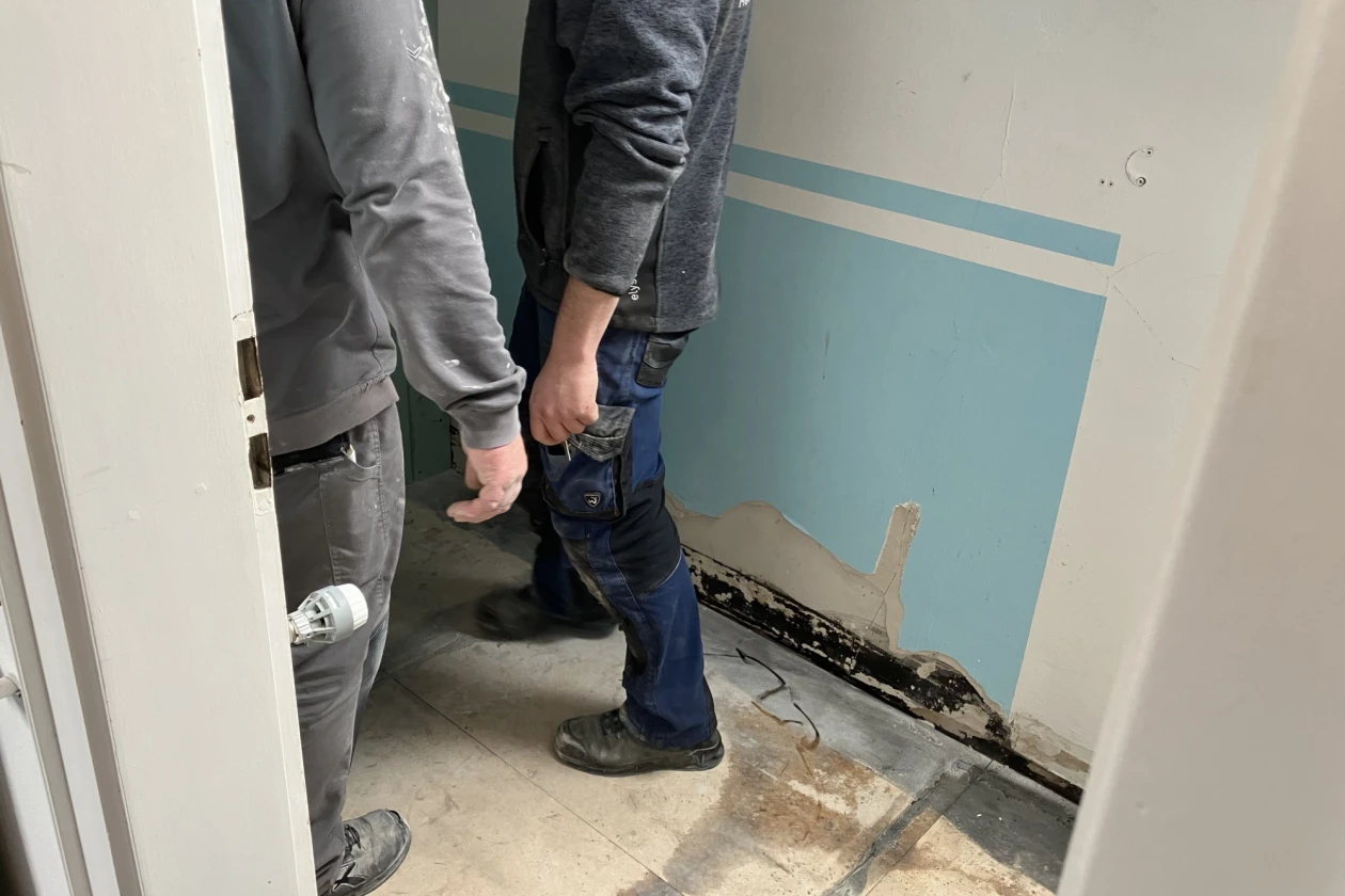 Zwei Handwerker stehen in einem Raum mit blauer Wand, wobei ein Wasserschaden im unteren Wandbereich sichtbar ist. Die Personen tragen graue Arbeitsbekleidung und scheinen die Sch&auml;den zu begutachten.