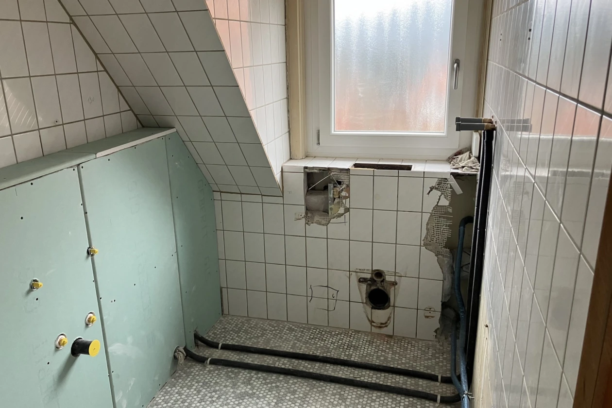 Schmales Badezimmer mit wei&szlig;en Wandfliesen und einem schr&auml;gen Dachgiebel. Toilettenhalterung und Installationen sind sichtbar, ein mattes Fenster l&auml;sst ged&auml;mpftes Licht herein.