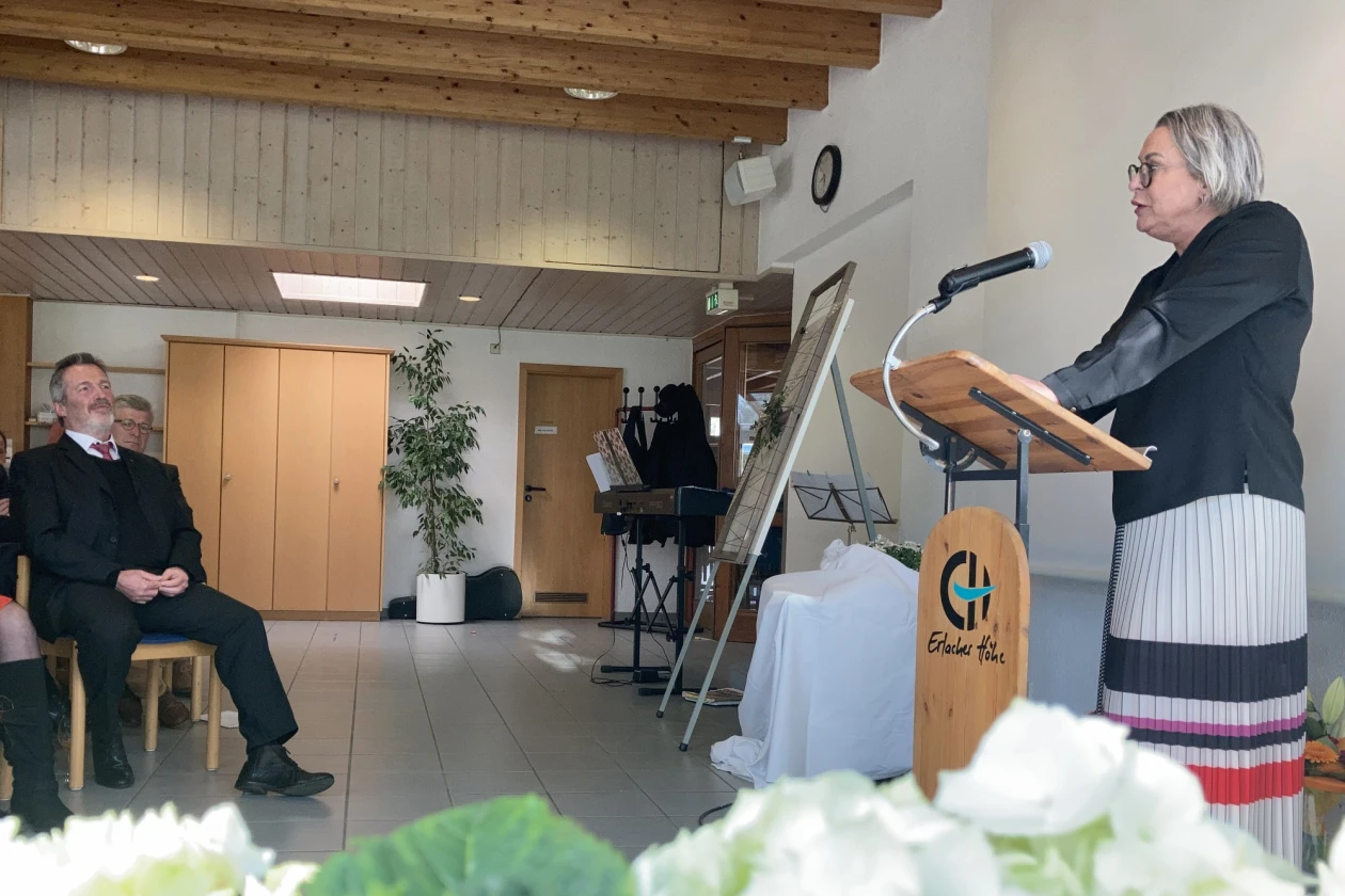 Eine Frau in kinnlangem grauen Haar und Brille, gekleidet mit einem schwarzen Oberteil und Strickrock steht an einem Rednerpult. Im Vordergrund sind Blumen zu sehen. Im Hintergrund sitzt ein dunkel gekleideter Mann mit grauem Haar und Bart, der zuh&ouml;rt.