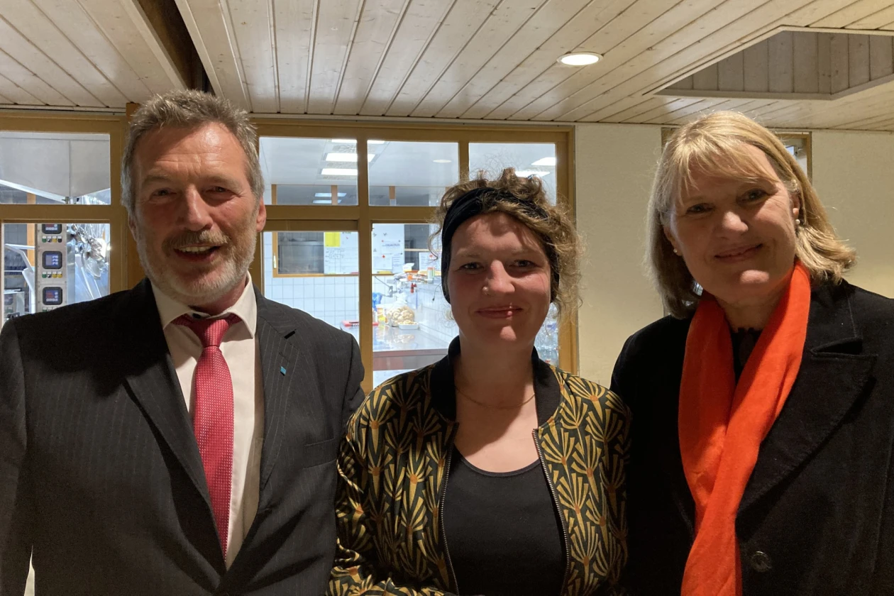 Mann mit grauem Haar und Bart, gekleidet mit schwarzem Jackett, wei&szlig;em Hemd und roter Krawatte neben einer jungen Frau mit gemusterter Jacke und schwarzem Shirt und einer weiteren Frau, schwarz gekleidet mit rotem Schal. Alle blicken freundlich l&auml;chelnd in die Kamera.