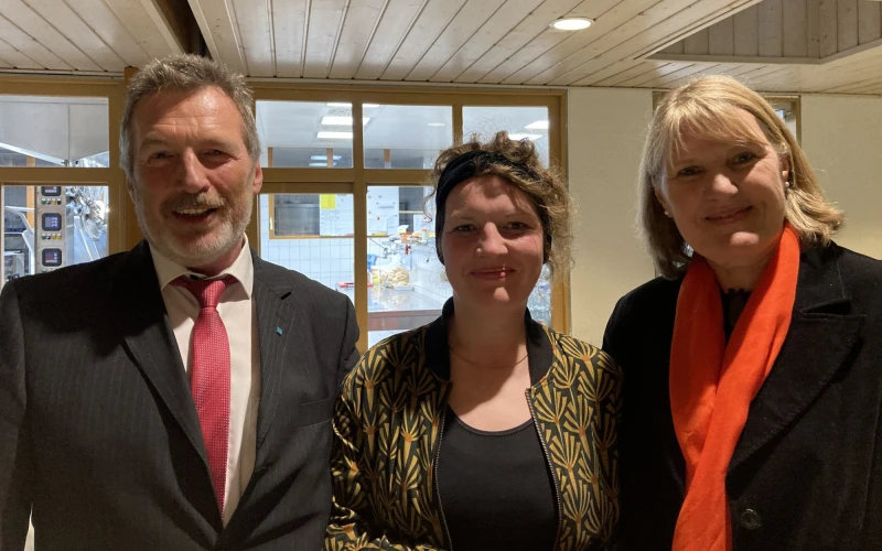 Mann mit grauem Haar und Bart, gekleidet mit schwarzem Jackett, wei&szlig;em Hemd und roter Krawatte neben einer jungen Frau mit gemusterter Jacke und schwarzem Shirt und einer weiteren Frau, schwarz gekleidet mit rotem Schal. Alle blicken freundlich l&auml;chelnd in die Kamera.