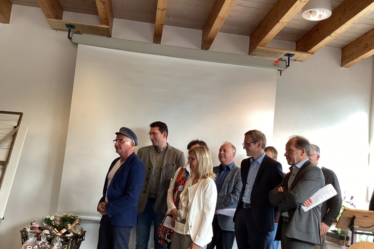 Eine Gruppe M&auml;nner und Frauen stehen vor einer wei&szlig;en Leinwand. Alle sind zur linken Seite gedreht und blicken in eine Richtung.