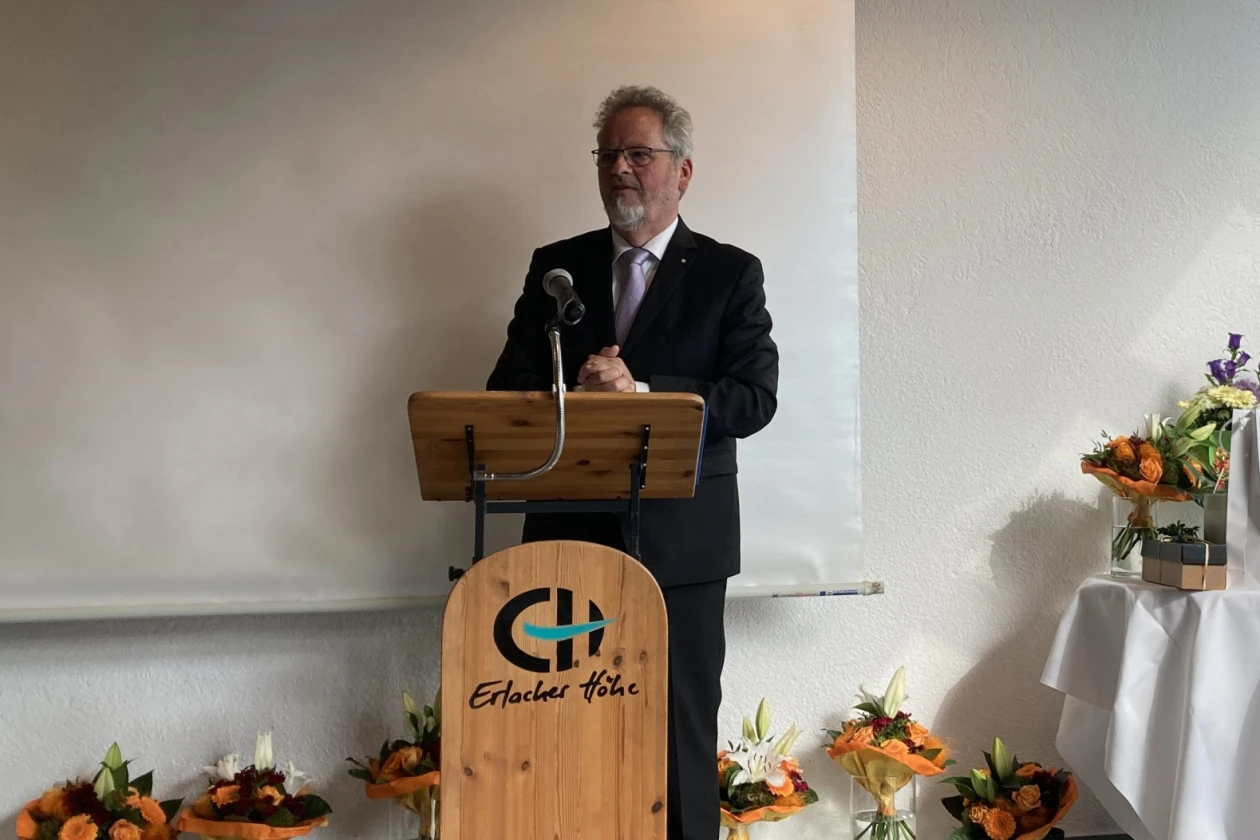 Mann mit lockigem grauen Haar und Bart steht an einem Rednerpult mit Aufschrift "Erlacher H&ouml;he" vor einer wei&szlig;en Leinwand. Im Hintergrund sind Blumenstr&auml;u&szlig;e und Geschenke zu sehen.