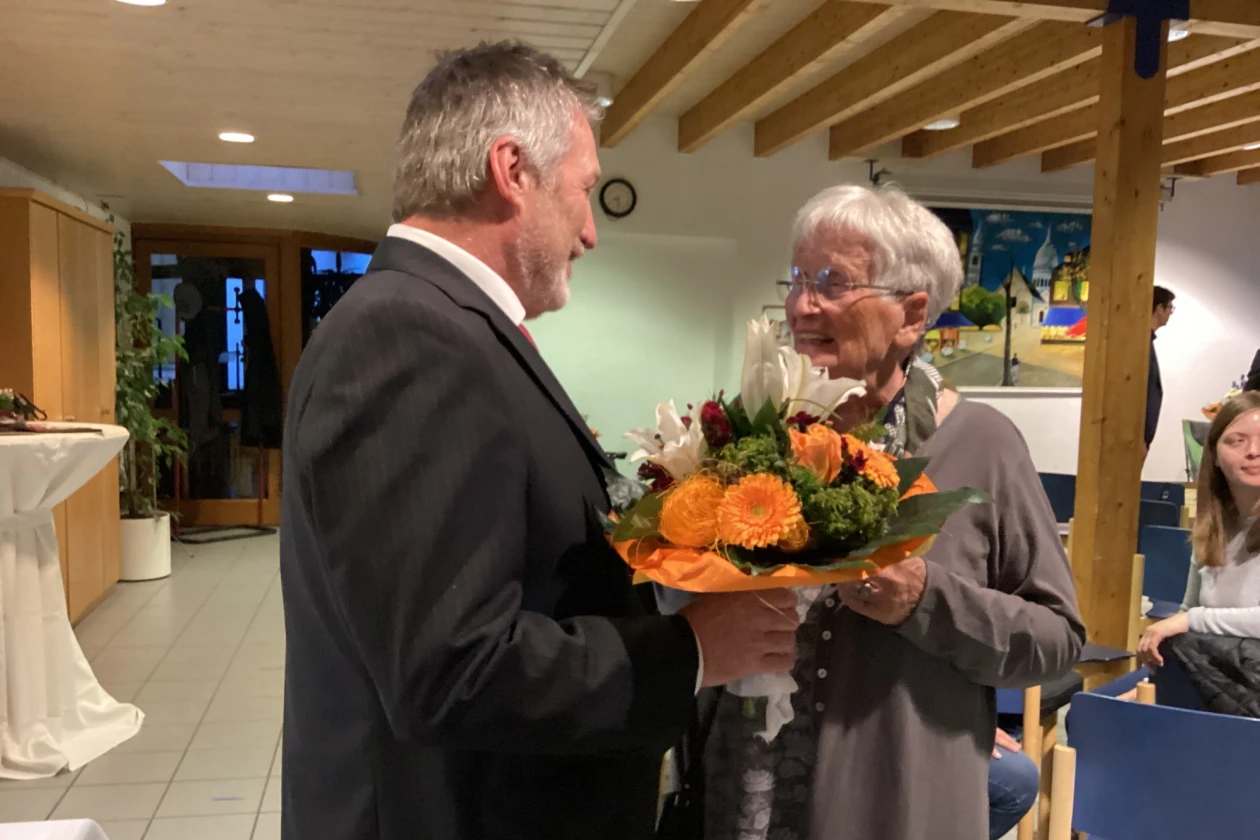 Mann mit grauem Haar und Bart steht mit einem Blumenstrau&szlig; vor einer &auml;lteren Frau mit grauem Haar. Beide l&auml;cheln sich an.