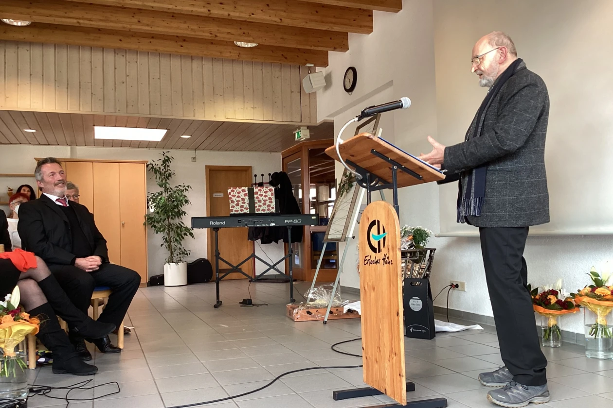 Ein &auml;lterer Mann mit dunklem Jackett und Schal steht vor einem Rednerpult. Vor ihm sitzt ein weiterer dunkel gekleideter Mann, der freundlich l&auml;chelt. Im Hintergrund sind weitere Personen zu sehen.