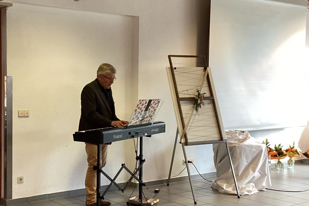 Mann mit schwarzem Jackett an einem E-Piano. Im Hintergrund eine Leinwand und ein Bilderrahmen auf einer Staffelei.