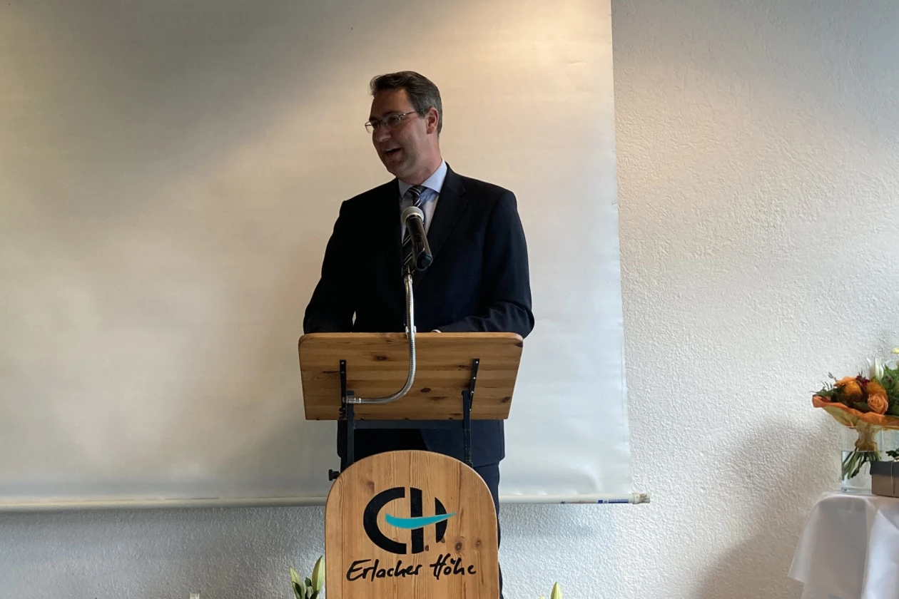 Mann mit dunklen Haaren, Brille und dunklem Anzug steht am Rednerpult mit der Aufschrift "Erlacher H&ouml;he". Im Hintergrund sind eine wei&szlig;e Leinwand und Blumenstr&auml;u&szlig;e zu sehen.