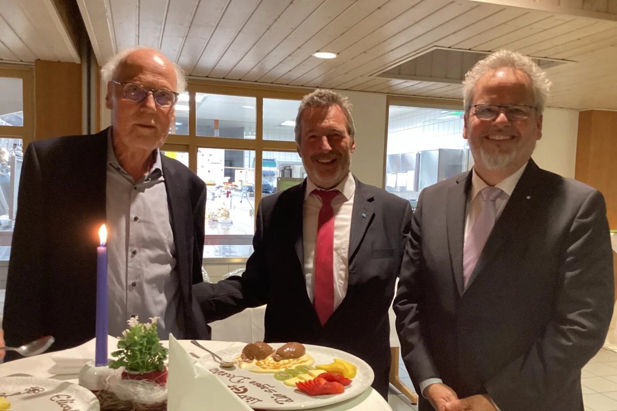 Drei Herren in Anz&uuml;gen stehen vor einem Stehtisch mit einer Kerze und Blumendekoration. Ein Teller mit Dessert steht vor ihnen. Alle drei blicken l&auml;chelnd in die Kamera.