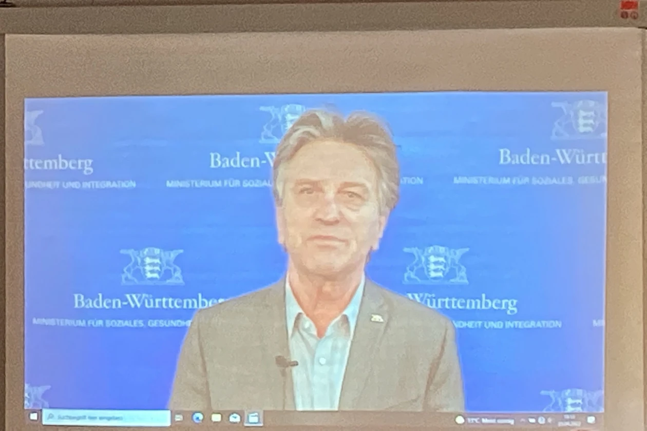 Auf einer Leinwand ist ein Mann mit hellgr&uuml;nem Hemd und grauen Jackett zu sehen. Er blickt in die Kamera. Er hat ein Mikrofon an seinem Jackett. Im Hintergrund das Wappen Baden-W&uuml;rttembergs.