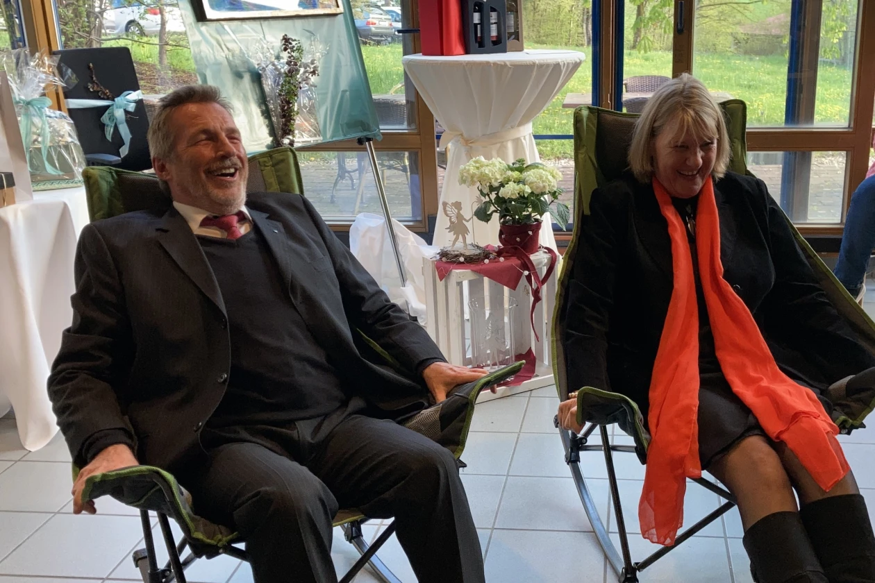 Mann mit grauem Haar und Bart in dunkler Kleidung in einem Campingschaukelsstuhl sitzend, daneben eine dunkel gekleidete Frau mit rotem Schal, ebenfalls in einem Campingschaukelstuhl. Im Hintergrund sind Blumen und Geschenke zu sehen.