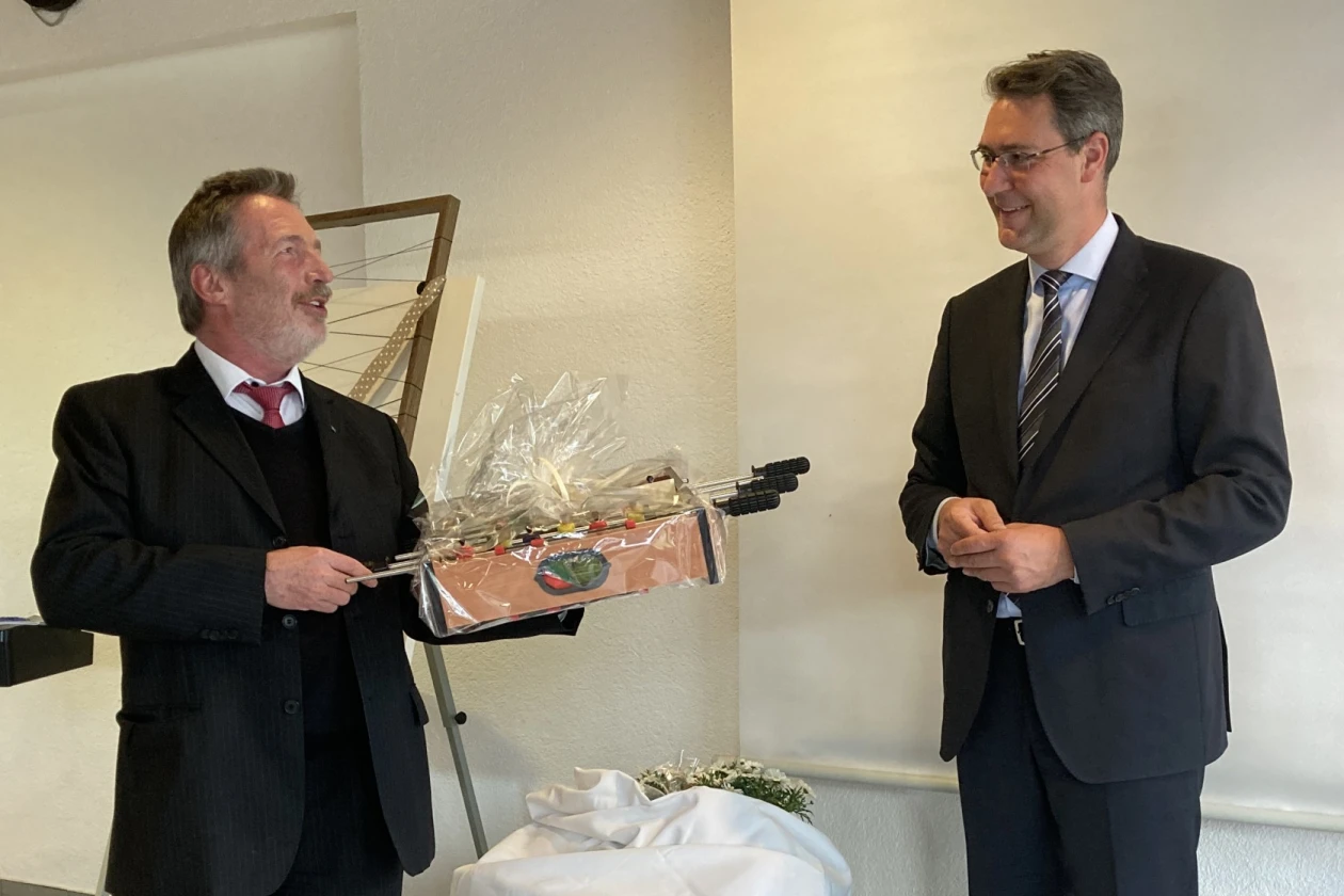 Mann mit grauen Haaren und Bart erh&auml;lt ein Geschenk von einem Mann mittleren Alters, beide in schwarzen Anz&uuml;gen gekleidet.