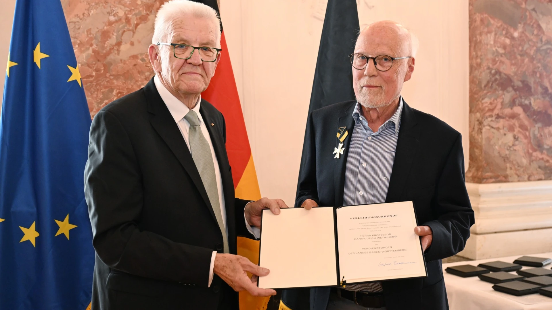 Zwei &auml;ltere M&auml;nner mit wei&szlig;em Haar mit einem Urkunden-Dokument. Ein Mann tr&auml;gt eine Medaille am Revers. Beide blicken in die Kamera. Im Hintergrund sind die Europa-, Deutschland- und Baden-W&uuml;rttemberg-Fahne zu sehen.